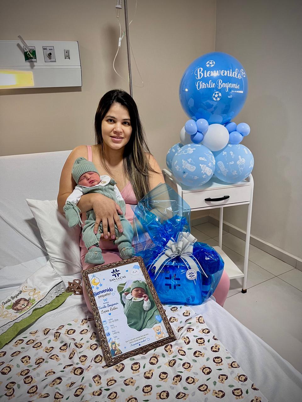 Mamá sonriente con su recién nacido en brazos en la habitación del Hospital San Lucas, junto al cuadro de bienvenida, el bolso de regalo y globos celestes de bienvenida